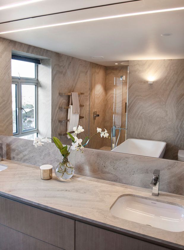 Contemporary bathroom interior viewed from the vanity, featuring a large stone-look benchtop with an integrated white basin, dark timber-toned vanity cabinetry, full-height warm grey-beige stone wall cladding, a frameless glass shower, freestanding bathtub, wall-mounted towel rail, recessed ceiling lights, and a large mirror. A window provides natural light, while an orchid arrangement adds a small decorative accent.