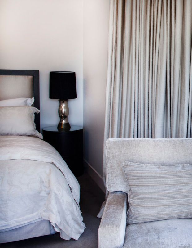 A softly styled bedroom corner with a partially visible upholstered bed, white patterned bedding, a black bedside table and lamp, full-height vertically striped curtains, and a plush light-grey armchair with a striped cushion. The palette is pale and neutral with dark contrasting furniture and flooring.