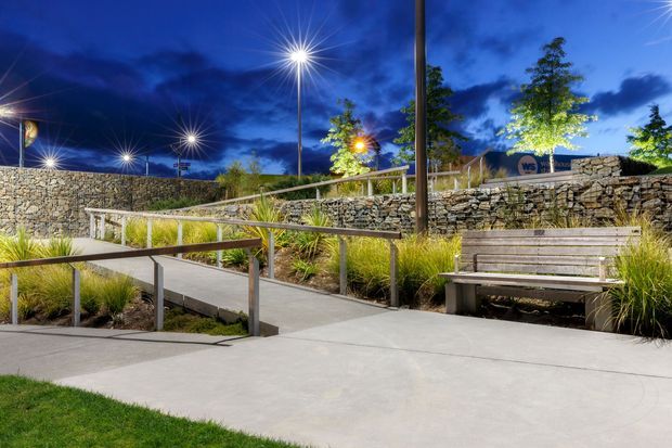 A landscaped outdoor public or commercial space at dusk, featuring accessible concrete pathways with metal handrails, timber bench seating, ornamental grasses and trees, and layered retaining walls made from stone-filled gabions. Tall contemporary lights illuminate the site against a deep blue evening sky.