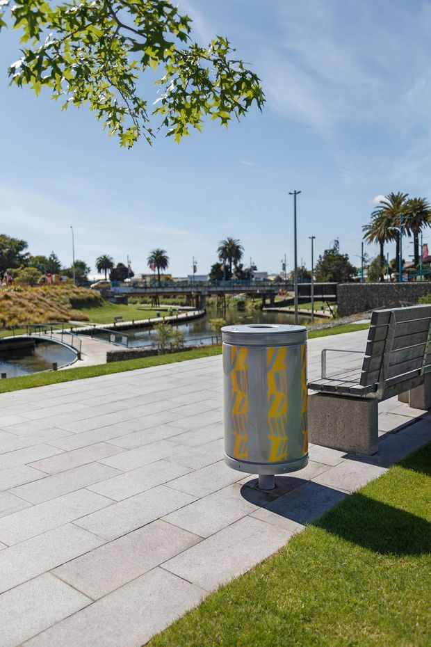 A sunny landscaped urban waterfront or canal-side public space with broad light-grey paved walkways, manicured grass, modern benches, and a cylindrical metal rubbish bin with yellow graphic markings in the foreground. A narrow water channel, small bridges, stone retaining walls, palm trees, and low-rise urban infrastructure are visible in the background.