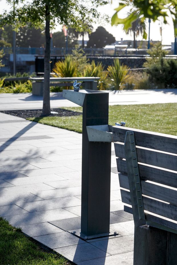 A landscaped public outdoor space with a modern timber-and-metal bench in the foreground, paired with a dark metal drinking fountain or water station. Pale rectangular paving, lawn, trees, ornamental grasses, and low planting surround the seating area. The scene is photographed in bright daylight with crisp shadows and a contemporary civic-landscape character.
