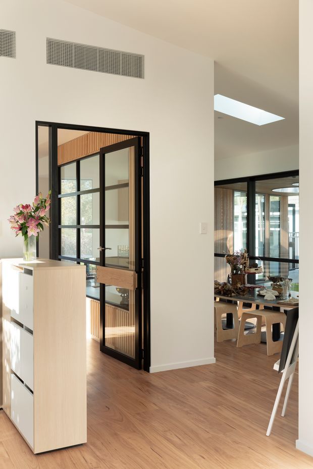 Bright contemporary interior hallway or entry area with warm timber flooring, white walls, a light timber storage console, and black-framed glazed doors. The glazed opening reveals a dining area with a timber table and chairs, vertical timber wall detailing, and abundant natural light. A skylight and linear ceiling vents add architectural interest.