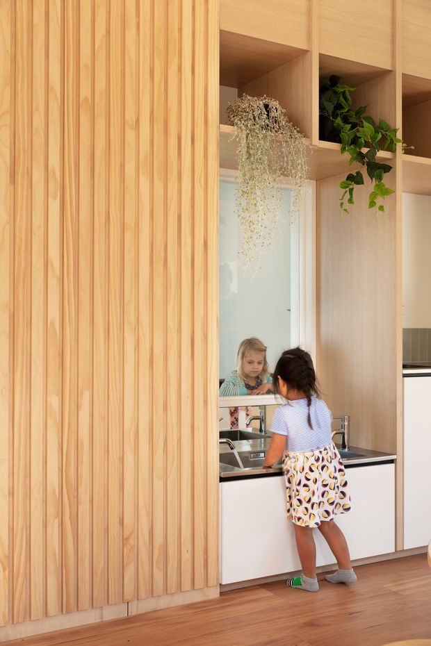 Warm contemporary kitchen interior featuring extensive light timber joinery. A vertical slatted timber feature wall occupies the left side, while open timber shelving with integrated planter niches frames a stainless-steel sink and black-edged benchtop. A large mirror or reflective splashback shows the child at the sink, and timber flooring continues through the space. The room is bright, natural, and family-oriented.