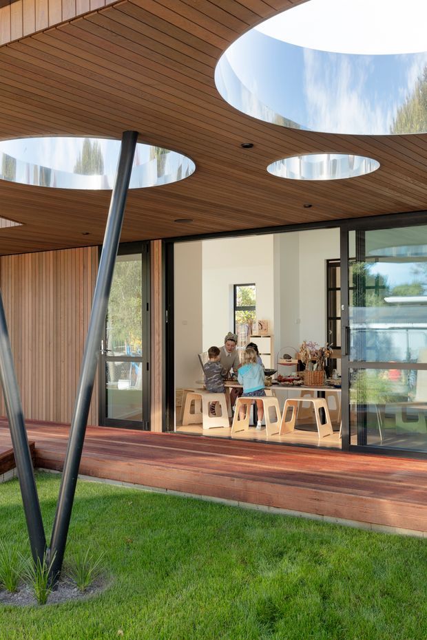 A modern single-storey residence viewed from a grassed garden, with a covered timber deck and a dramatic curved timber-lined canopy pierced by several circular skylight openings. Slender angled steel posts support the canopy, while expansive black-framed glazed sliding doors reveal a bright dining and kitchen area with children seated at a table. The design combines strong indoor-outdoor connection, warm timber surfaces, and sculptural organic roof geometry.
