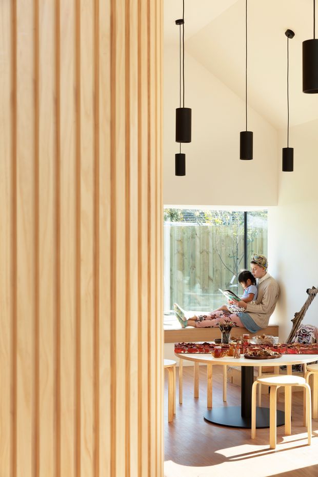 Bright contemporary interior with a tall vertical timber slat feature wall in the foreground, a warm timber floor, and a dining area beyond. Several black cylindrical pendant lights hang from the high angled ceiling. A round light-coloured dining table with stools and benches occupies the room, with a large window bringing in sunlight and views of a timber fence. The space feels warm, airy, and family-oriented.