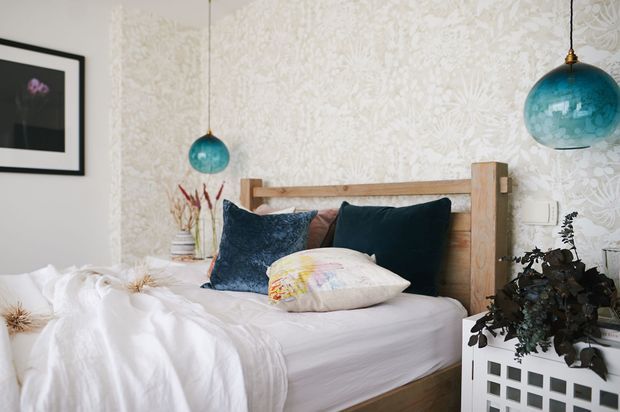 A cozy bedroom with a natural timber bed frame, white bedding, dark teal and patterned cushions, and botanical wallpaper. Two turquoise glass pendant lights hang beside the bed, with framed artwork and a plant-filled white bedside cabinet adding decorative detail.