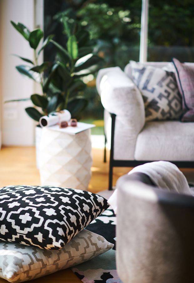 A warmly lit contemporary living room vignette featuring a pale upholstered sofa and armchair, patterned black-and-white floor cushions, a faceted white side table, indoor greenery, a large window, and light timber flooring. The room has a bold monochrome textile pattern balanced by soft neutral upholstery and natural greenery.