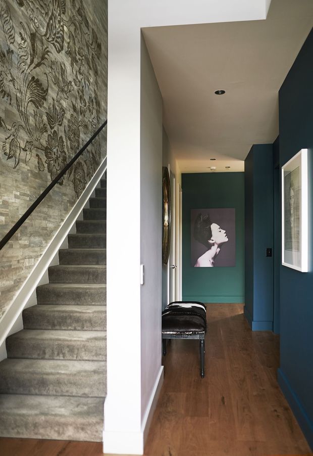 A narrow residential hallway beside a carpeted staircase. The corridor has deep blue-green painted walls and cabinetry-like trim, warm timber flooring, recessed ceiling lights, a framed portrait, and a black upholstered bench. The stair wall features patterned wallpaper above a stacked-stone finish, with a dark handrail and light trim.
