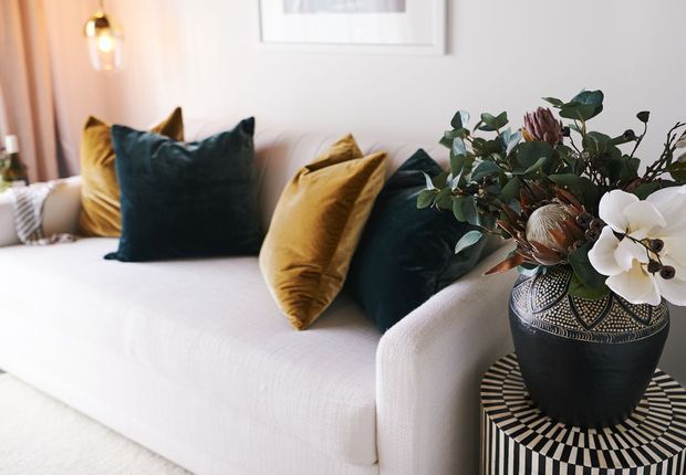 A close-up of a refined living room with a white upholstered sofa, deep teal and mustard cushions, a dark patterned vase with botanical stems on a round striped side table, soft neutral walls, framed artwork, and warm pendant lighting.