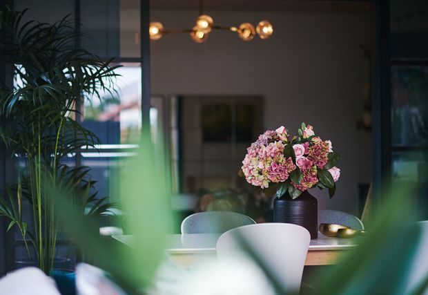 A softly lit contemporary dining area viewed through lush indoor plants. A round dining table with pale upholstered chairs is centred beneath a sculptural globe pendant light, with a large floral arrangement as the focal point. Glazing and dark-framed architectural elements create a moody, refined atmosphere.