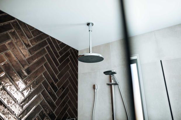 Close-up of a contemporary bathroom shower area, featuring a ceiling-mounted round rain shower, handheld shower fitting, pale large-format wall tiles, and a dark glossy herringbone tile feature wall. A black-framed shower screen is visible at the right.