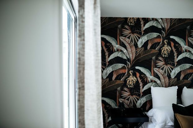 A close view of a bedroom corner featuring a bed with white bedding, a black bedside table, and a dramatic dark botanical feature wallpaper with large tropical leaves and flowers. A tall window with a light curtain provides soft natural light.