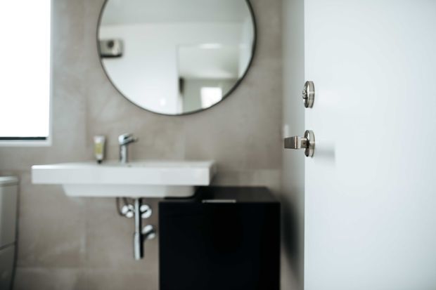 Compact contemporary bathroom with a wall-mounted white rectangular basin, chrome tapware and exposed plumbing, a black vanity cabinet, beige stone-look wall tiles, and a large round mirror. A partially open white door and window are visible.