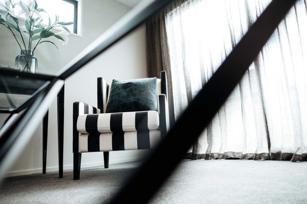 A softly lit interior corner featuring a modern upholstered accent armchair with bold black-and-cream vertical striping and a dark teal cushion. The chair sits on a light grey carpet beside sheer floor-to-ceiling curtains, with a leafy plant on a dark glass-topped side table. Dark diagonal foreground elements partially frame the view.