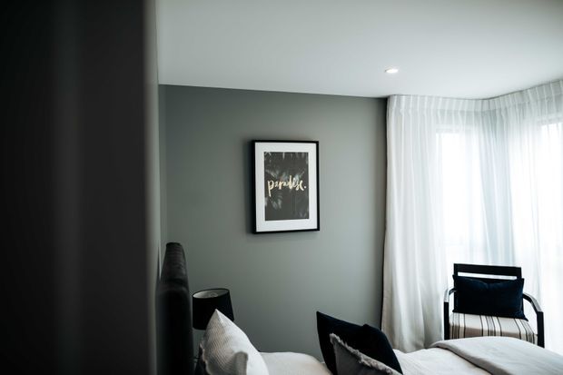 A softly lit bedroom with muted grey-green walls, sheer white floor-to-ceiling curtains wrapping around a corner window, a framed artwork, bedside lamp, dark accent chair, and the edge of a white bed with textured cushions. The room has a calm, contemporary feel with clean lines and restrained styling.