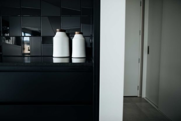 A close-up interior view of a dark, minimalist built-in joinery area with glossy black tiled wall cladding and a black stone-look countertop or shelf. Two white ceramic vessels sit on the surface, while a white-painted hallway or doorway with grey flooring is visible to the right.