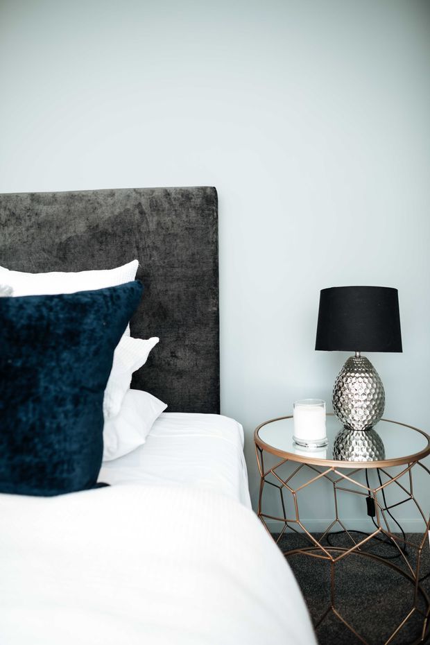 A close-up of a contemporary bedroom corner featuring a bed with crisp white bedding, a dark charcoal upholstered headboard, and a deep navy accent cushion. Beside the bed is a round glass-topped side table with a geometric copper-toned metal base, topped by a reflective silver textured lamp with a black shade and a clear drinking glass. The room has pale blue-grey walls and dark carpeting.