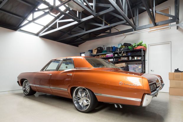 A polished orange classic car dominates a spacious garage or workshop. The room has a smooth light-coloured concrete floor, plain white walls, exposed dark steel roof trusses, high-level glazing or skylights, open storage shelving with tools and equipment, and stacked cardboard boxes. The industrial structure and vehicle-focused fit-out are clearly visible.