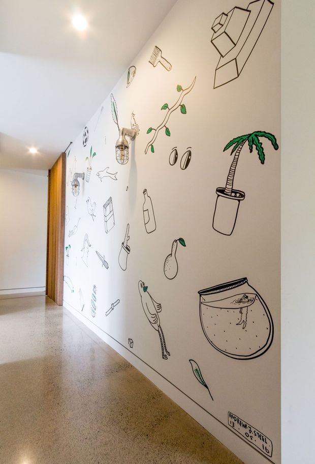 A bright interior hallway with polished concrete flooring and white walls covered in playful black-and-green hand-drawn illustrations, including plants, animals, objects, and a fishbowl. Timber detailing frames an opening at the end of the corridor, while recessed ceiling lights provide even illumination.
