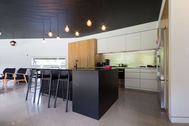 Open-plan contemporary kitchen and dining area with a central black island, white flat-panel cabinetry, timber pantry joinery, pale tiled flooring, and a dark ceiling with multiple suspended pendant lights. A dining table and chairs are visible in the adjoining area.