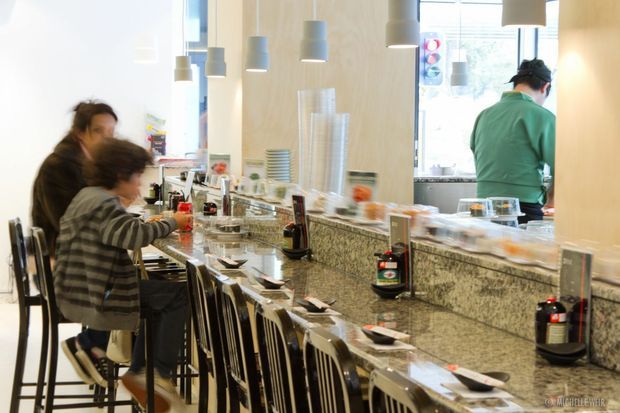 Bright contemporary counter-service dining space with a long speckled stone counter, rows of high stools, pendant lights, table settings, and large windows. Diners sit at the counter while staff work behind it.