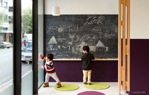 A compact children's creative nook beside full-height glazing, featuring a large wall-mounted chalkboard covered in children's drawings, a dark purple lower wall, pale upper wall, timber vertical screen at the right, pendant lighting, and two children drawing and playing on round green mats.