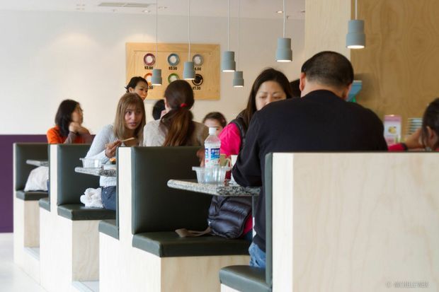 A bright casual dining interior with customers seated in booth-style tables. The space features pale timber booth partitions and tables, dark upholstered seating, a light neutral wall, colourful pendant lights, and a small wall-mounted display. The image appears to show a café or restaurant fit-out.