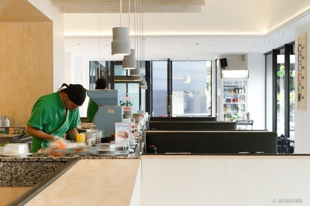 Bright contemporary café interior viewed from the customer area toward the service counter and open preparation kitchen. The space features pale timber cabinetry and ceiling elements, white surfaces, dark-framed glazing, pendant lights, refrigerated drink display, and black counter fronts. Staff are working behind the counter, with outdoor greenery visible through the windows.