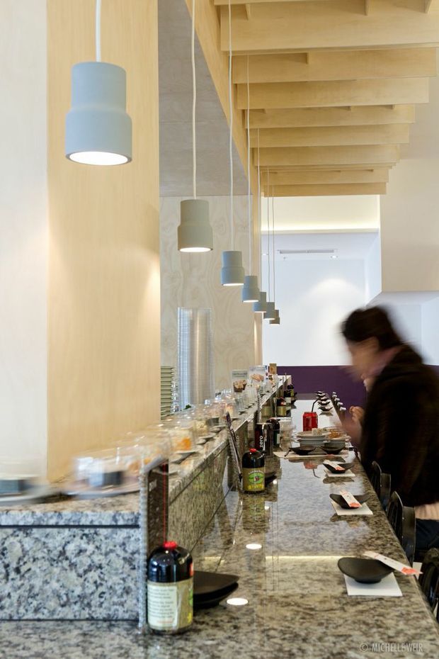 A narrow contemporary restaurant or bar interior with a long polished stone counter lined with bowls, plates, bottles, and table settings. Pale timber wall panels and a dramatic exposed timber slatted ceiling structure run through the space, while repeated cylindrical pendant lights hang above the counter. The palette is light timber, pale stone, white, and muted purple, with a blurred person at the service counter.