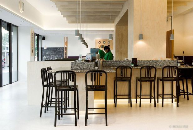 A bright contemporary commercial café or hospitality interior with a long stone-topped bar and numerous black timber bar stools. The space features a pale polished floor, white walls, light timber ceiling beams and wall cladding, pendant lighting, dark-framed glazing, and a partially visible service counter and kitchen area.