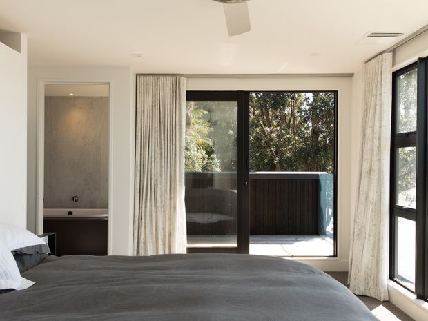 Bright contemporary bedroom with a large grey bed in the foreground, a partially visible ensuite bathroom with a freestanding or built-in bath at left, and full-height black-framed glazing opening to a private outdoor terrace. White walls and ceiling, pale curtains, and abundant natural light create a calm, refined interior.