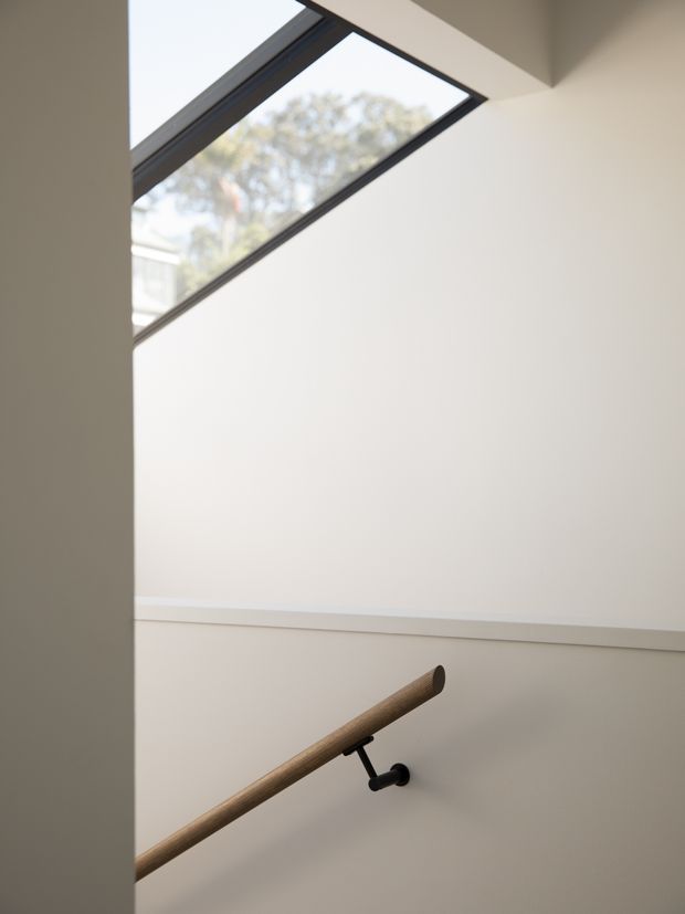 A minimalist stairway viewed from the side, featuring a warm timber handrail mounted on dark metal brackets, smooth white walls, and a large angled skylight or clerestory window bringing in natural light. The space is clean, restrained, and contemporary, with a view of trees outside through the glazing.