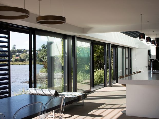 A bright, contemporary open-plan interior with a dining area and kitchen connected by extensive floor-to-ceiling glazing. Dark-framed sliding glass doors overlook a body of water and landscaped greenery. The space features a pale smooth ceiling with recessed detailing, light timber flooring, a white kitchen island, dark dining furniture, and suspended circular and pendant lighting.