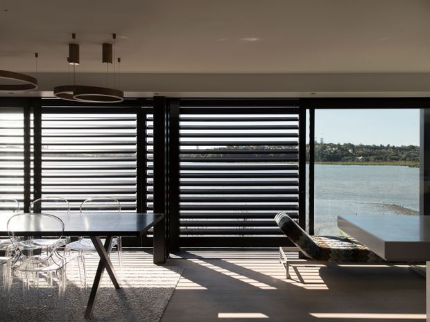 A contemporary waterfront dining and living area with expansive glazing, dark horizontal louvres, and strong linear sunlight. The space includes a dining table with transparent chairs, sculptural ring pendant lights, and a low lounge/daybed beside the window. Calm water and a distant shoreline are visible outside.
