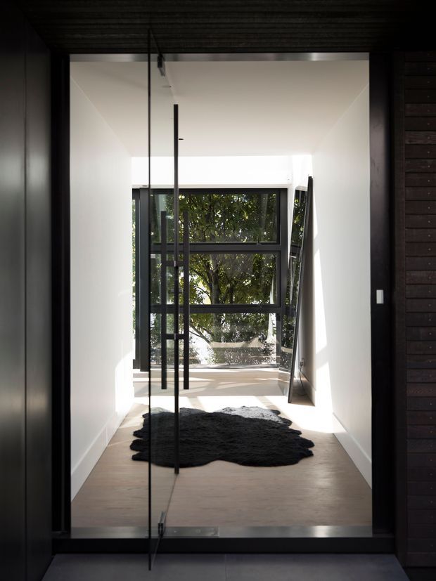 A minimalist interior hallway or entry passage framed by dark timber and glazing. White walls and ceiling lead toward large black-framed windows overlooking leafy greenery, with a pale smooth floor and a dark irregular-shaped rug. A tall, slim black-framed glass door or screen stands in the foreground, creating a strong linear composition and contrast between the bright corridor and dark surrounding timber surfaces.