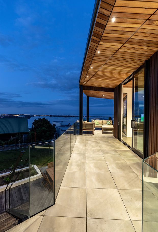 Twilight view of a contemporary upper-level terrace or balcony with large-format pale stone-look tiled flooring, frameless glass balustrades, dark slender structural posts, and extensive floor-to-ceiling glazing. A warm timber-lined soffit with recessed downlights extends over the terrace, leading toward a furnished outdoor lounge and an interior living area. The balcony overlooks a residential waterfront or coastal setting with distant lights.