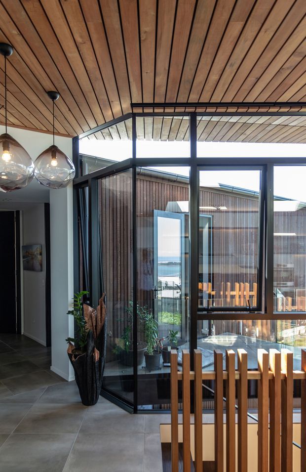 Interior view of a modern open-plan circulation and dining area with a warm timber slatted ceiling, dark metal-framed floor-to-ceiling glazing, and views through the courtyard toward a waterfront. Sculptural glass pendant lights hang above the dining area, while timber vertical slats form a balustrade in the foreground. Large-format grey floor tiles, indoor plants, and timber-clad exterior elements are visible.
