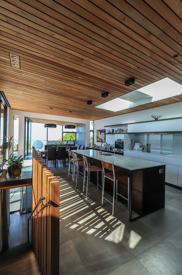 A bright, open-plan kitchen and dining area with a long dark island and integrated bar seating, white built-in cabinetry, extensive glazing, and a timber slatted ceiling. A timber slatted stair or balustrade element occupies the foreground, while two large ceiling skylights bring in strong sunlight and cast linear shadows across the tiled floor. The space has a clean, refined contemporary character with warm timber detailing.
