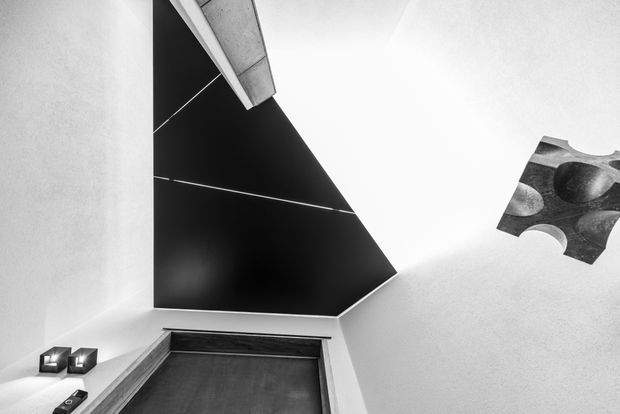 Monochrome architectural interior detail showing a dramatic angular black feature wall or ceiling plane intersecting white plastered surfaces. A narrow concrete-toned structural element projects near the top, while a dark framed doorway or opening occupies the lower portion. Small geometric wall-mounted fixtures and an abstract monochrome artwork add sparse visual detail. The composition is highly graphic, minimalist, and strongly contrasted.