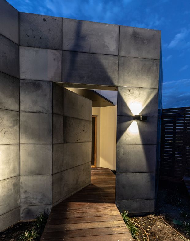 Dusk exterior view of a contemporary entry, framed by large weathered concrete panels. A timber boardwalk leads through a recessed doorway, with warm interior light and a geometric up-and-down wall sconce creating a dramatic focal point. A dark horizontal-slat privacy fence is visible at right against the blue evening sky.