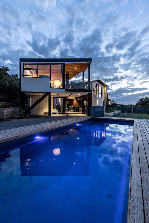 A contemporary multi-level house viewed from the pool at dusk. The home has a strong geometric composition with dark metal framing, large glazed openings, white rendered walls, and warm timber soffits and ceilings. A timber pool deck surrounds a deep blue illuminated swimming pool, which reflects the house and dramatic cloudy sky. Covered outdoor living areas and an upper balcony are visible.