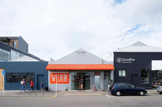 Street-level view of a contemporary low-rise commercial precinct with dark painted facades, white gabled roof forms, and a prominent orange-red corrugated awning over the Welder entrance. The adjacent GoodFor storefront has a dark facade and white branding. Urban street frontage with parked cars and pedestrians.