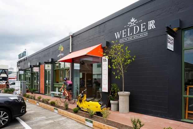 Street-level exterior view of a contemporary commercial retail strip with dark painted brick facades, large glazed shopfronts, bright orange awnings, signage, planter boxes, and a paved pedestrian edge. The image includes visible wellness and hospitality-style businesses, with urban streetscape context and parked vehicles in the foreground.
