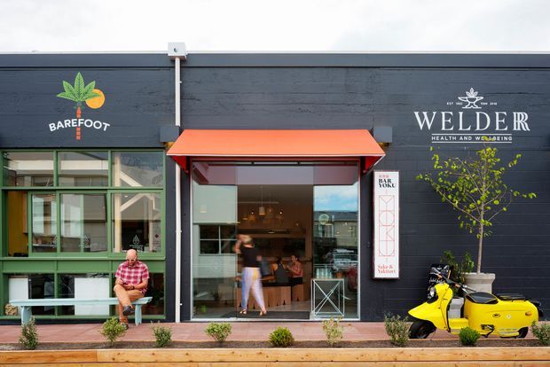 Street-facing view of a low-rise commercial building with dark horizontal timber cladding, green-framed glazing, and a bright red-orange awning over an open café or restaurant entrance. Visible signage identifies Barefoot Health and Welder Hair and Beauty, with a yellow scooter parked outside, planter boxes, and small shrubs along the pedestrian frontage.