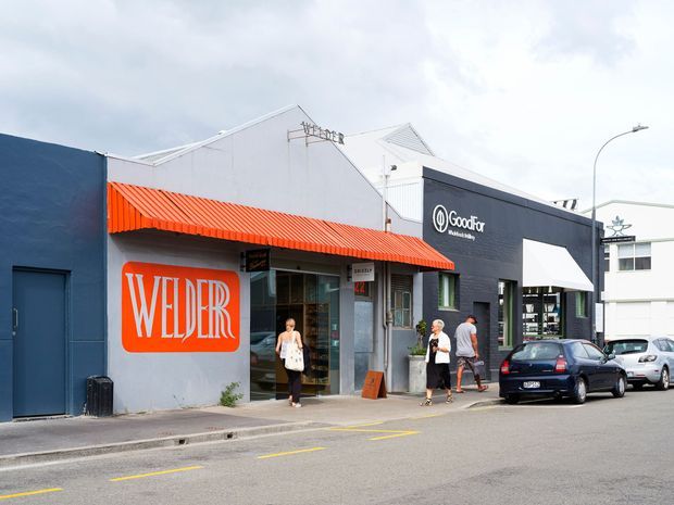 Street-level view of a row of low-rise commercial shopfronts in an urban setting. The foreground building has a dark blue facade, a bright orange corrugated awning, and a large orange Welder sign. Adjacent premises include a dark charcoal Goodfor storefront and a white commercial building, with parked cars and pedestrians along the street.