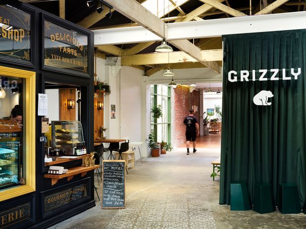 Interior view of a converted industrial-style commercial food hall or market. A dark, traditional-style bakery or pastry storefront with gold lettering and display cases occupies the left, while a deep green curtained shopfront marked “GRIZZLY” is on the right. The shared passage has patterned concrete or tile flooring, exposed timber roof trusses, white structural beams, pendant lighting, potted plants, and brick detailing in the distance.