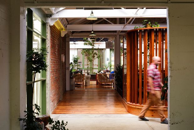 A warm, adaptive commercial interior viewed through a broad opening, with a timber-floored passage leading to a casual seating area. Exposed red brick walls, painted structural beams and ceiling elements, industrial pendant lights, large glazed openings, and a curved screen of vertical stained timber battens define the space. Indoor plants and wicker seating add an organic touch, while a blurred person is walking through the foreground.