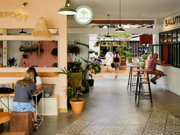 A bright, casual hospitality interior, likely a café or food-and-beverage venue. The open-plan space features a patterned concrete floor, pale plaster walls, exposed ceiling services and structure, hanging pendant lights, timber and metal bar seating, indoor plants, wall-mounted shelving, and a service counter. Customers are seated at a window bench and along a high communal counter, with an open courtyard or outdoor dining area visible beyond.