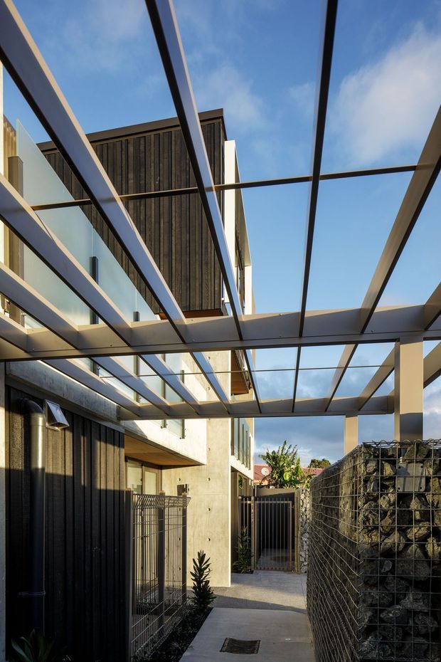 A narrow contemporary exterior walkway or courtyard between modern buildings, viewed upward and along a paved path. The space features a large steel-framed glass pergola or covered canopy, dark vertical timber cladding, exposed concrete walls, metal gates and fencing, and a gabion stone retaining wall. Clear blue sky is visible through the canopy, with sparse planting along the edges.