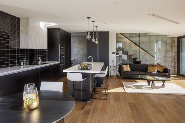 Open-plan contemporary kitchen and living area with dark cabinetry, a central island, black tiled splashback, timber flooring, concrete-look walls, pendant lighting, and a glass-balustraded staircase. A dining table occupies the foreground, while the living zone includes a sofa and coffee table.
